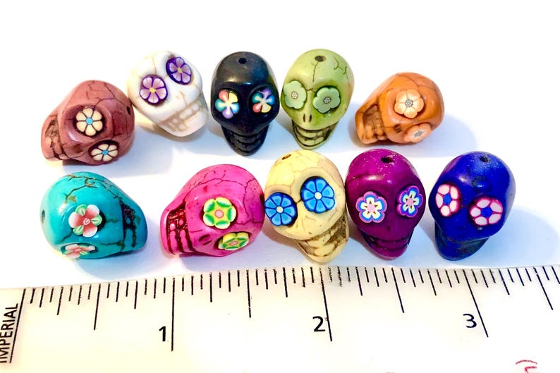 May include: Ten colorful sugar skull beads with floral designs. The beads are approximately 1 inch in diameter and are arranged on a ruler.
