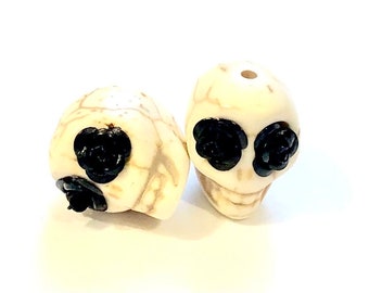 May include: Two white ceramic skull beads with black rose accents. The beads are approximately 2.5 cm in diameter and have a hole drilled through the top for stringing.