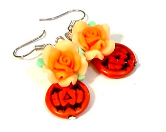 May include: A pair of Halloween-themed earrings. Each earring features a small orange pumpkin charm with a carved face, paired with a peach-colored rose and green accents. The earrings have silver-colored hooks.