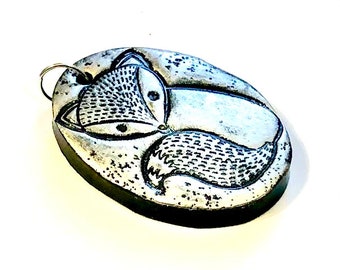 May include: A silver oval pendant with a black and white fox design. The pendant has a silver loop for attaching a chain.