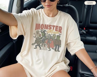 May include: Ivory-colored t-shirt featuring the text "MONSTER Mash" above a cartoon illustration of classic Halloween monsters. The graphic includes a mummy, witch, werewolf, vampire, Frankenstein's monster, and a ghost. The shirt has short sleeves.