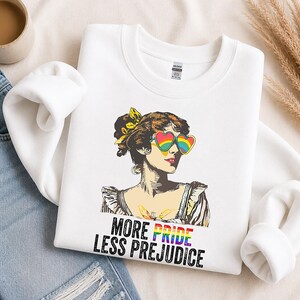 May include: A white crewneck sweatshirt featuring a graphic of a person with an updo hairstyle wearing heart-shaped rainbow sunglasses. Below the illustration, the text reads MORE PRIDE LESS PREJUDICE, with the word PRIDE in rainbow colors.