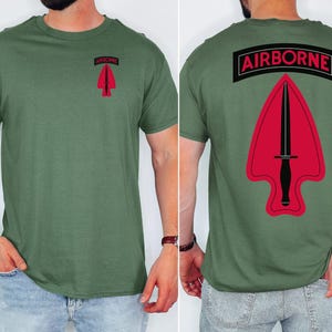 Puede incluir: Camiseta verde oliva con un gráfico rojo y negro en la parte delantera y trasera. El gráfico frontal es una flecha roja con un cuchillo negro, y el gráfico trasero es una versión más grande del mismo diseño con la palabra "AIRBORNE" encima.