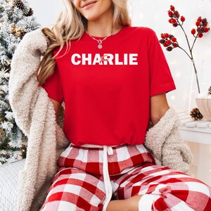 A woman wearing a red t-shirt with the name "CHARLIE" and red and white plaid pajama pants is sitting in front of a Christmas tree.
