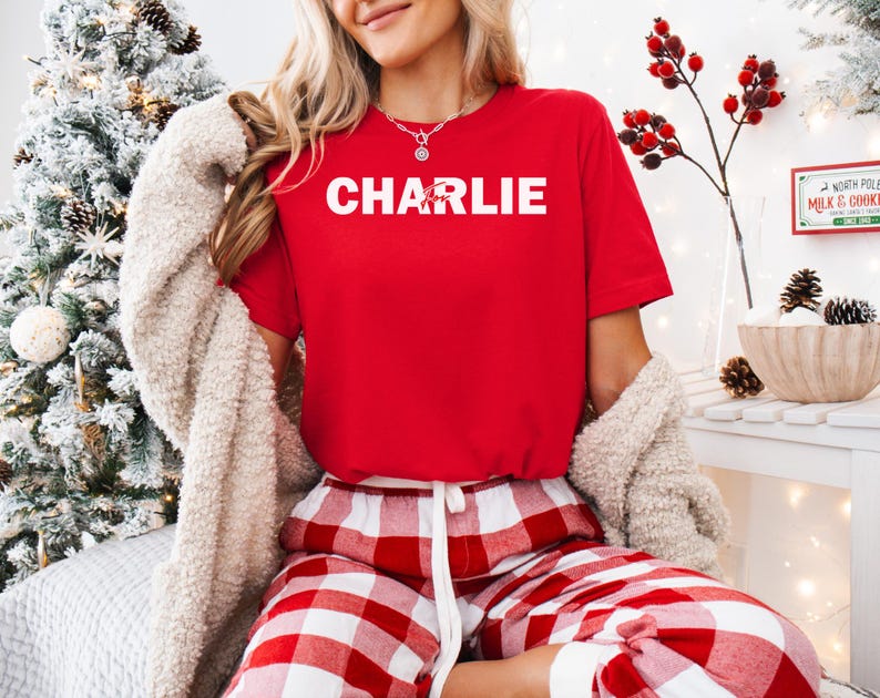 A woman wearing a red t-shirt with the name "CHARLIE" and red and white plaid pajama pants is sitting in front of a Christmas tree.