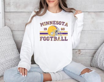 May include: White long-sleeve sweatshirt with a graphic design. The design features the text "MINNESOTA FOOTBALL 1960" in purple and gold, with a football helmet graphic. The sweatshirt is worn by a person.