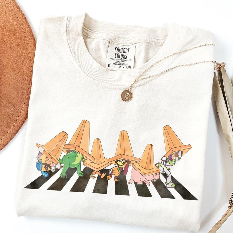 May include: Cream-colored Comfort Colors t-shirt featuring a cartoon graphic of characters crossing a crosswalk, each wearing an orange traffic cone. A gold necklace with a small pendant rests on the shirt.