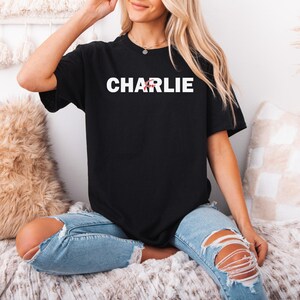 A woman wearing a black t-shirt with the word "CHARLIE" printed on it, sitting on a bed with a beige blanket and a pillow.