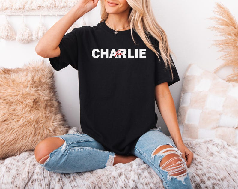 A woman wearing a black t-shirt with the word "CHARLIE" printed on it, sitting on a bed with a beige blanket and a pillow.