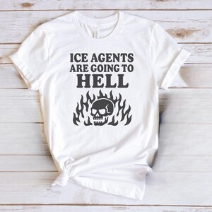 May include: White t-shirt with the text "ICE AGENTS ARE GOING TO HELL" in black, above a black skull graphic surrounded by flames. The shirt is knotted at the bottom.