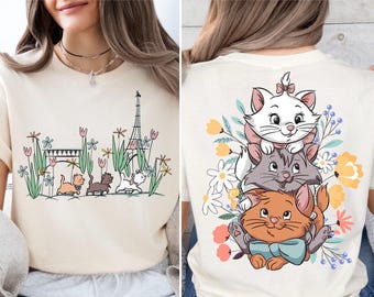 May include: Cream-colored t-shirt featuring a graphic of three cartoon kittens stacked on top of each other, surrounded by flowers. The front of the shirt has a graphic of kittens, flowers, and the Eiffel Tower.