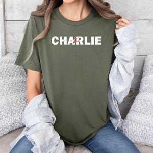 A person wearing a green t-shirt with the name "CHARLIE" printed on it, sitting on a couch with a gray sweater draped over their arm.