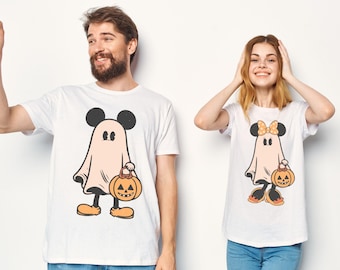May include: Two white t-shirts featuring graphic prints of cartoon characters dressed as ghosts for Halloween. The left shirt shows a character with round ears holding an orange jack-o'-lantern, and the right shirt shows a character with a polka-dot bow.