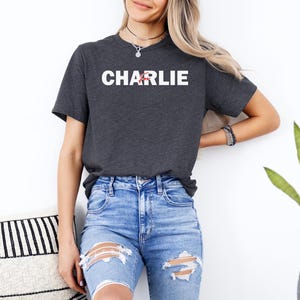 A woman wearing a grey t-shirt with the word "CHARLIE" printed on it, paired with distressed blue jeans, stands in front of a white wall.