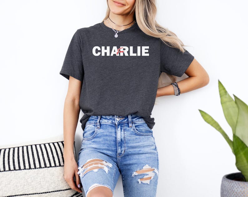 A woman wearing a grey t-shirt with the word "CHARLIE" printed on it, paired with distressed blue jeans, stands in front of a white wall.