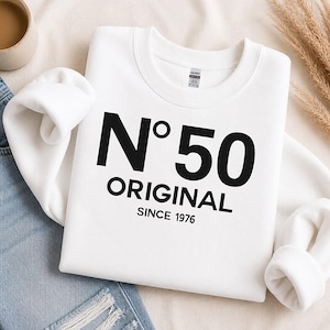 May include: A white crewneck sweatshirt featuring black text that reads N°50 ORIGINAL SINCE 1976. The garment has long sleeves and a ribbed collar, cuffs, and hem. The design is centered on the chest in a bold, sans-serif typeface.