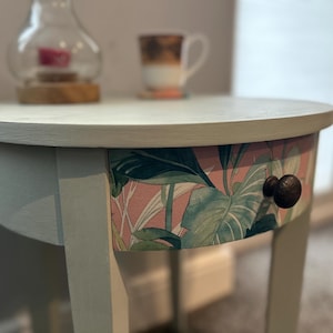 May include: A round, light blue side table with a single drawer. The drawer front is covered in a pink and green floral patterned paper. The drawer pull is a dark brown knob.
