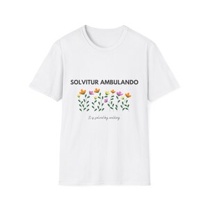 Può includere: Maglietta bianca con una stampa grafica di fiori e il testo "SOLVITUR AMBULANDO" e "It is solved by walking."