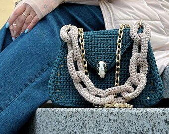 May include: A teal blue crocheted handbag with a gold chain strap and a snake-shaped clasp. The bag is sitting on a gray stone surface.