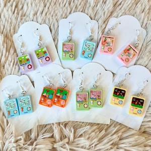 Può includere: Un set di 10 paia di orecchini pendenti con console per videogiochi in miniatura con grafica pixelata colorata. Gli orecchini sono esposti su cartoncino bianco.