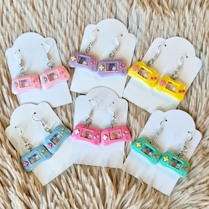 May include: Six pairs of dangle earrings featuring a miniature handheld game console design. The earrings are displayed on white card stock with a silver hook. The consoles are in various colors, including pink, purple, yellow, blue, and green.