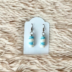 May include: A pair of milk carton earrings with blue tops and the word "MILK" printed on the front. The earrings are hanging from a white card.