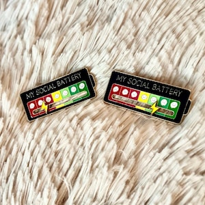 Puede incluir: Dos pines de esmalte negro y dorado con forma de batería con un medidor de batería social. El medidor está lleno de caras sonrientes amarillas, caras fruncidas rojas y caras neutrales verdes. El texto "MY SOCIAL BATTERY" está impreso en cada pin.