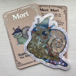May include: A holographic sticker of a cartoon snail wearing a hat, with a matching card that says "Mort" and "Regard The Hero". The sticker has a shiny, iridescent finish. The card has text that says "Meet Mort".