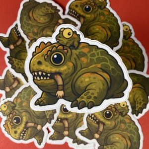 Puede incluir: Una pegatina troquelada con una ilustración de dibujos animados de una criatura grande, verde y verrugosa parecida a un sapo con un sapo más pequeño en la cabeza. La criatura tiene ojos grandes y oscuros, dientes afilados y sostiene una mano.