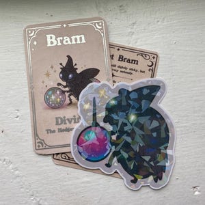 May include: A sticker and two cards featuring a whimsical illustration. The sticker depicts a dark silhouette of a creature holding a crystal ball. The cards have the name "Bram" and the words "Divine The Hedgehog".