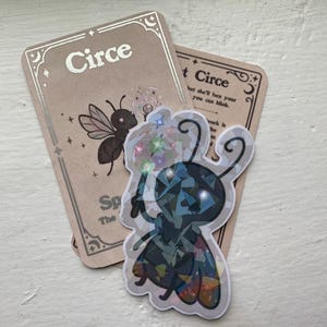 May include: A holographic insect sticker holding a wand, placed over two cards. The cards feature the word "Circe" and an insect illustration. The cards have a beige background and decorative borders.