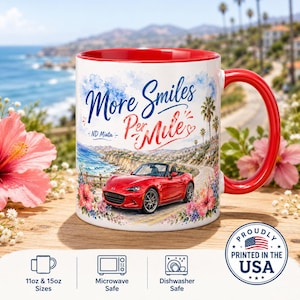 May include: A white ceramic mug with a red interior and handle, featuring a red convertible car on a coastal road. The text reads More Smiles Per Mile and ND Miata. The design includes watercolour-style flowers and palm trees on a white background.