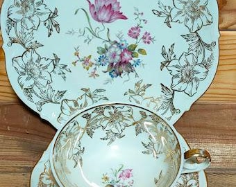 Può includere: Un servizio da tè in porcellana composto da un piatto grande, un piattino e una tazza. Ogni pezzo presenta uno sfondo azzurro pallido, contorni floreali dorati e un bouquet centrale di fiori rosa, viola e blu con un manico dorato.