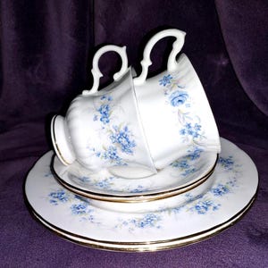 May include: A white teacup and saucer set with a delicate blue floral pattern. The set includes a teacup, a saucer, and a smaller plate, all with gold trim. The teacup is tilted, showcasing the intricate design.