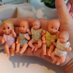 Könnte beinhalten: Fünf kleine Plastikpuppen, die Babys darstellen, werden in einer Hand gehalten. Die Puppen haben bemalte Gesichtszüge und tragen verschiedenfarbige Outfits. Die Outfits sind rosa, blau, gelb und weiß.