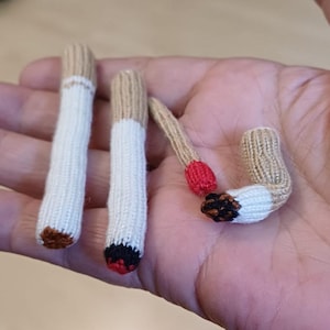 Può includere: Sigarette lavorate a maglia a mano in varie fasi di completamento. Le sigarette sono bianche con filtri beige e estremità marroni o nere. Una sigaretta ha la punta rossa e un'altra è parzialmente fumata, con cenere nera e marrone.