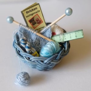May include: A blue wicker basket filled with blue yarn, knitting needles, and a small book titled "Stricken leichtgemacht".