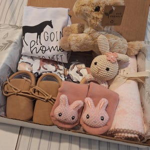Op de afbeelding: Een geschenkdoos met een wit t-shirt met een zwarte paardenafbeelding en de tekst GOIN home TO MEET, een beige pluche paard, een gehaakte konijnenrammelaar, beige babymocassins, roze sokken met konijnengezichtjes en een gevouwen roze deken.