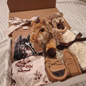 Può includere: Una scatola regalo contenente un body per neonati color marrone chiaro con grafica di cavallo, una fascia abbinata, un paio di mocassini, un peluche a forma di cavallo con etichetta ecologica, un sonaglio a orsetto e una coperta avvolgente.