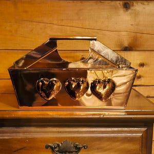 May include: A copper-colored metal basket with a handle and three heart-shaped cutouts on the front, sitting on a wooden nightstand. The nightstand has a drawer and two doors with dark metal handles. The background is a wooden wall.