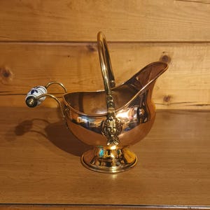 May include: A polished copper coal scuttle with a brass handle and a pedestal base. The vessel features a decorative brass lion head accent where the handle attaches and a side handle with a white ceramic grip decorated with blue floral patterns.