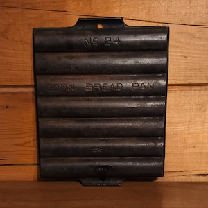 May include: A dark, rectangular cast iron cornbread pan with a handle. The pan has a series of parallel, rounded ridges. The words "NO 24" and "CORN BREAD PAN" are embossed on the pan.