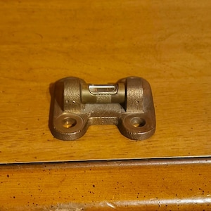 May include: A small, antique brass level is mounted on a wooden surface. The level has a bubble indicator and two mounting holes. The wood has a warm, medium tone with visible grain. The image also shows a drawer with a decorative handle.