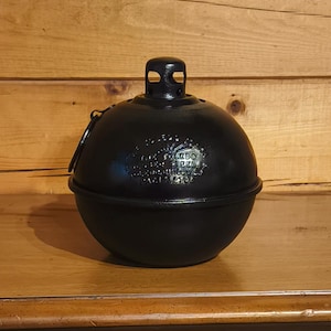 May include: A black, spherical oil lamp with a handle and a top, resting on a wooden nightstand. The lamp has embossed text on its surface. The nightstand is made of wood and has two drawers and two doors.