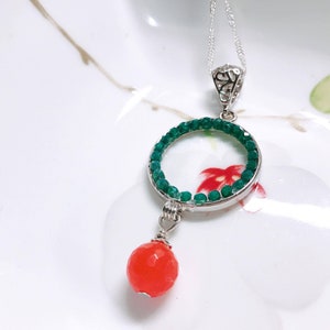 May include: A silver necklace with a circular pendant. The pendant features a ring of green beads and a dangling orange bead. The necklace is displayed on a white surface with floral accents.