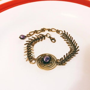 May include: A gold-toned bracelet with a purple flower charm and a chain made of small, pointed links. The bracelet is on a white surface.