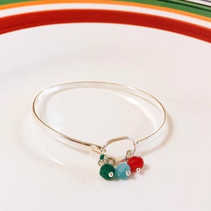 Puede incluir: Un brazalete de plata con un dije que tiene tres pequeñas cuentas de piedras preciosas. Las cuentas son rojas, azules y verdes.