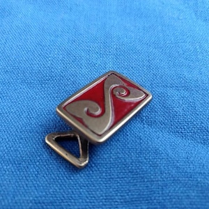 May include: A silver-toned metal belt buckle with a red enamel inlay featuring a stylized design. The buckle has a triangular loop for attaching to a belt.