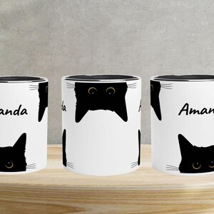 May include: White ceramic mug with a black rim and handle, featuring a repeating pattern of black cat faces and the name "Amanda" in black script. The mug is displayed on a wooden surface.