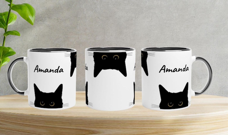May include: White ceramic mug with a black rim and handle, featuring a repeating pattern of black cat faces and the name "Amanda" in black script. The mug is displayed on a wooden surface.